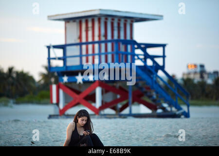 Eine Dame saß am South Beach in Miami SMS mit Start und Streifen Strandhütte hinter Ihnen Stockfoto