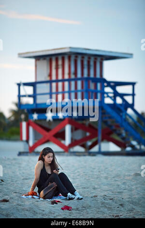 Eine Dame saß am South Beach in Miami SMS mit Start und Streifen Strandhütte hinter Ihnen Stockfoto