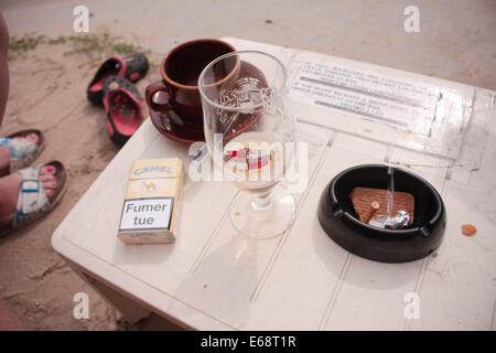 Ein Bier, Kaffeetasse, Schachtel Zigaretten und Aschenbecher auf einem weißen Tisch außerhalb Stockfoto