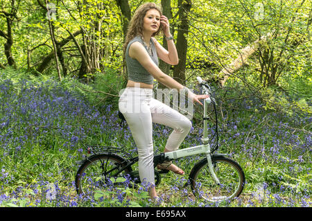 (In Richtung Kamera) ein hübsches junges Mädchen sitzt auf einem Fahrrad unter Bluebells in den Wäldern bei Banstead, Surrey, England, UK. Stockfoto