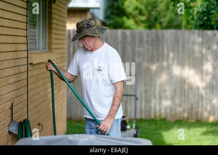 Ein 76-jähriger Kaukasier erledigt Hofarbeiten, indem er draußen einen Wasserschlauch gegen das Haus wickelt. USA Stockfoto