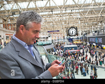 Reife Geschäftsmann mit einem iPhone in London Waterloo Station mit belebten Bahnhofshalle im Hintergrund Stockfoto