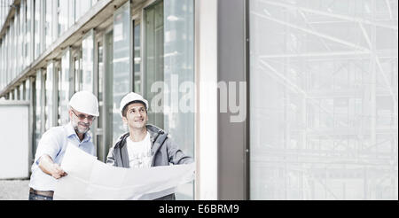 Zwei Ingenieure, 55-65 und 25-35 Jahre alt, planen harte Hüte tragen, stehen vor einem Gebäude und Blick auf ein Gebäude Stockfoto