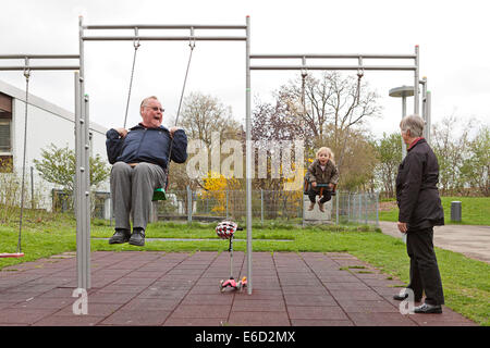 Großvater und seine Enkelin, 3 Jahre, auf Schaukeln, Großmutter stehen neben ihnen spielen Stockfoto