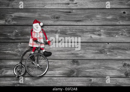 Lustige rote Weihnachtsmann auf hölzernen grauen Hintergrund in Eile für den Kauf von Weihnachten präsentiert auf einem alten Fahrrad sitzen. Stockfoto