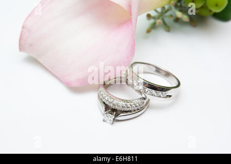 Weißgold und Diamanten Trauringe und einen Verlobungsring sind auf einem weißen Hintergrund neben Rosa Hochzeitsblumen angezeigt. Stockfoto