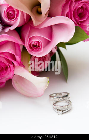 Weißgold und Diamanten Trauringe und einen Verlobungsring sind auf einem weißen Hintergrund neben Rosa Hochzeitsblumen angezeigt. Stockfoto
