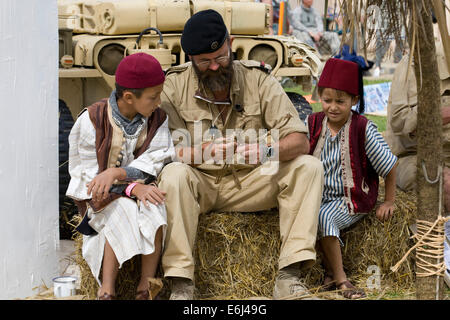 Soldat macht eine Schilfpuppe mit traditionell gekleideten Kindern, die zuschauen Weiter auf dem Lager der Wüstenratten in Kandahar Stockfoto