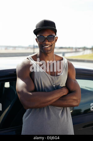 Porträt von hübschen afrikanischen Kerl mit seinen Armen gekreuzt an einem Auto lächelnd gelehnt. Stilisierte muskulösen Kerl mit Sonnenbrille. Stockfoto