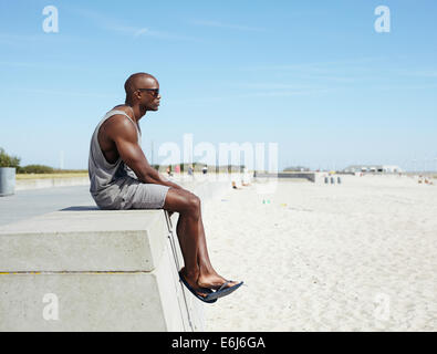 Seitenansicht der jungen afrikanischen Mann sitzt auf einer Strandpromenade wegsehen. Afro amerikanische Modell entspannen im Freien am Strand mit co Stockfoto