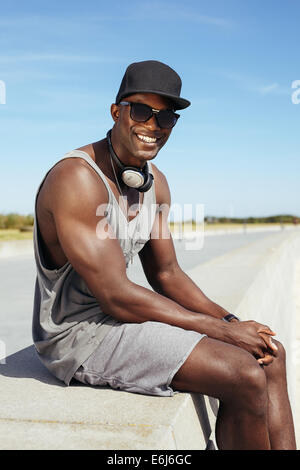 Porträt von glücklicher junge afrikanische Mann sitzt auf einer Promenade. Stilvolle Männermodel trägt Sonnenbrille und Kappe. Stockfoto