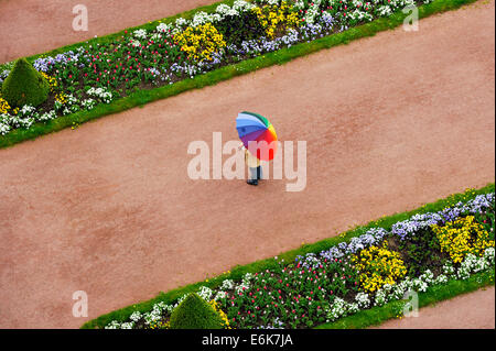 Person mit Schirm in Regenbogenfarben auf einem roten Kies Weg zwischen Blumenbeeten im Schlossgarten Stockfoto