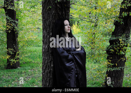 Frau in schwarz stehen gegen einen dunklen Baumstamm in den grünen herbstlichen Wald Stockfoto