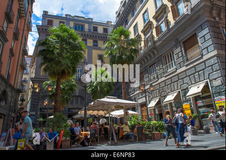 Touristen und Einheimische sitzen in einem externen Café oder Restaurant mit einem reich verzierten Uhr und Palmen in Neapel, Italien. Stockfoto