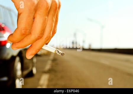 Nahaufnahme eines Mannes mit einer brennenden Zigarette in der Hand und wartet neben einem Auto parkte neben einer nicht befahrenen Straße Stockfoto