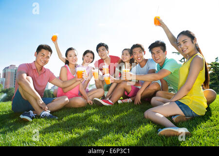 Fröhliche junge Erwachsene halten Saft sitzen auf Rasen Stockfoto