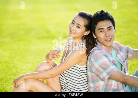 Fröhliches junges Paar sitzen Rücken an Rücken auf dem Rasen, Musik hören Stockfoto