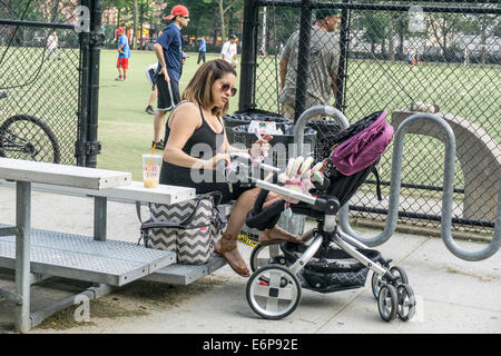 dralle glücklich ziemlich junge Mutter sitzt in Tribüne & versucht, ihr kleines Baby im Kinderwagen zu amüsieren, wie Mann Softball New York spielt Stockfoto