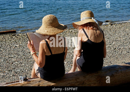 Zwei pensionierte Frauen sitzen auf einem log Bücher lesen am Strand, Bowen Island, BC, Kanada Stockfoto