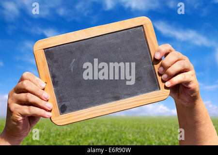 Menschliche Hände halten eine Schiefer Tafel mit leeren Werbeflächen in der Luft im Hintergrund einer grünen Wiese mit blauem Himmel Stockfoto