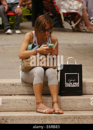 Eine junge Frau sitzt auf einem Schritt in die Sonne, Blick auf ihr Handy mit Einkaufstasche in Venedig Stockfoto