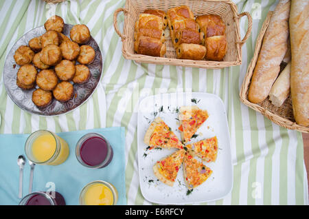 Picknick-Frühstück mit Saft, Gebäck, Croissants, Brotkorb, Tischdecken, Geschirr, Baguettes auf einem Tisch verteilen Stockfoto