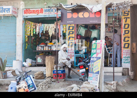 Typische Straßenszene in einem Dorf in der Nähe von Deogarh Rajasthan: Blick auf ein ladenbesitzer mit weißen Turban draußen sitzen kleine lokale Geschäfte Stockfoto
