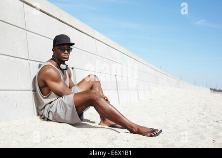 Hübscher Kerl mit Kopfhörer und Sonnenbrille am Strand neben einer Wand sitzen. Muskuläre afro-amerikanische Männermodel entspannend. Stockfoto