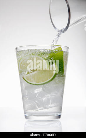 Clear water pouring from jug into drinking glass with lime and ice cubes Stockfoto