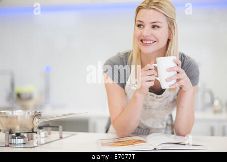 Junge Frau Kaffee trinken und mit Blick auf Rezeptbuch in Küche Stockfoto