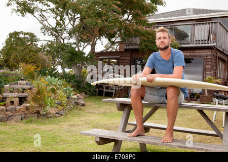 Porträt von männlichen Surfer auf Picknickbank mit Surfbrett auf Schoß sitzen Stockfoto