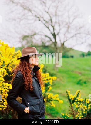 Eine Frau mit langen lockigen Haaren, trägt einen Hut, von einem blühenden Ginster Strauch. Stockfoto