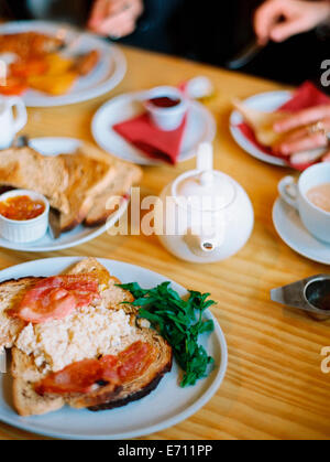 Ein Tisch mit Platten von Lebensmitteln gedeckt. Ein warmes Frühstück mit Speck, Eiern und Toast und Tee. Zwei Personen sitzen Stockfoto