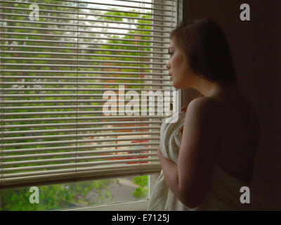 Mürrisch junge Frau blickt aus Hotel Schlafzimmerfenster während in Blatt gewickelt Stockfoto
