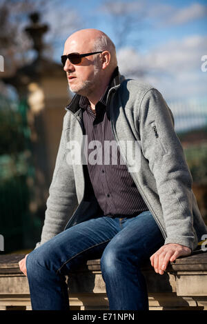 Glatze Mann mittleren Alters in Jeans sitzen im Freien in der Sonne auf einer Treppe eine Zeitung lesen Stockfoto