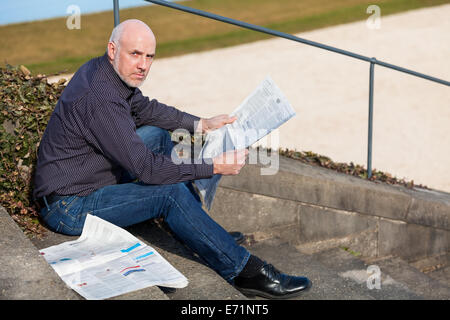 Glatze Mann mittleren Alters in Jeans sitzen im Freien in der Sonne auf einer Treppe eine Zeitung lesen Stockfoto