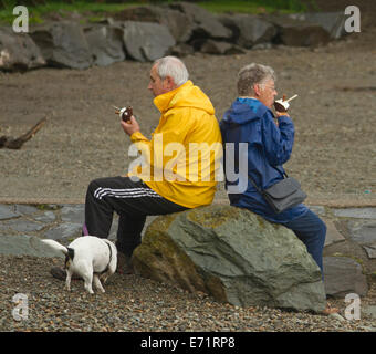 Älteres Ehepaar tragen Regenmäntel sitzt auf Felsen essen Eis neben Coniston Water im Lake District, Cumbria, England Stockfoto