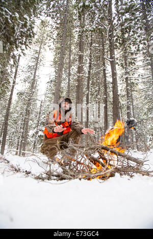 Ein männlicher Jäger bleibt neben einem Feuer im Schnee warm. Stockfoto