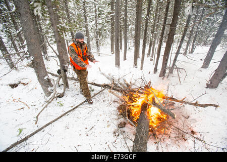 Ein männlicher Jäger bleibt neben einem Feuer im Schnee warm. Stockfoto