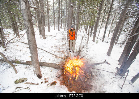 Ein männlicher Jäger bleibt neben einem Feuer im Schnee warm. Stockfoto