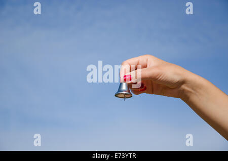 Hand mit roten Nägeln schütteln kleine silberne Glocke und Jingle sound auf Hintergrund des blauen Himmels. Stockfoto