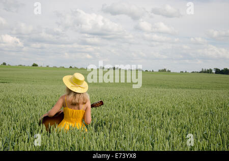 Land-Frau im gelben Kleid und Hut auf dem Kopf spielen mit Gitarre im Weizenfeld. Stockfoto