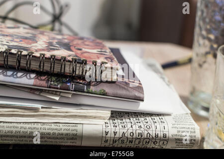 Ein Stapel von Zeitschriften, Zeitungen und Büchern auf einem Tisch neben Trinkgläsern. Das fertige Kreuzworträtsel ist kopfüber sichtbar. Stockfoto