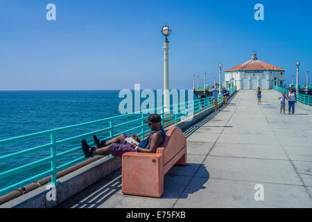 Ein afroamerikanischer männliche Touristen, liest ein Buch auf einer Bank und andere gehen auf die kommunalen Pier in Manhattan Beach, Kalifornien. Stockfoto