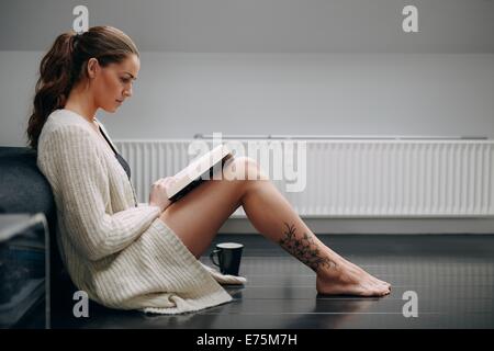 Seitlicher Blick auf schöne junge Dame ein Buch zu lesen. Kaukasisches weibliches Modell zu Hause sitzen am Boden durch eine Couch mit einem Roman. Stockfoto