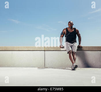 Bild des Fit Jüngling entspannend auf Damm nach seinem Lauf gegen blauen Himmel. Afrikanische Sportler im Freien mit Exemplar. Stockfoto