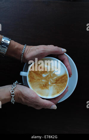 Reife Frau in Café hält eine Tasse Kaffee Cappuccino über Kopf anzeigen Stockfoto