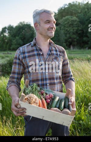 Lächelnd reifen Mann tragen Kiste von erntefrischem Gemüse im Garten Stockfoto