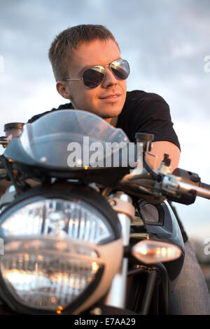 Romantische Portrait schöne Biker Mann in Sonnenbrille sitzt auf einem Fahrrad auf einen Sonnenuntergang in der Nähe von See und Stadt Stockfoto