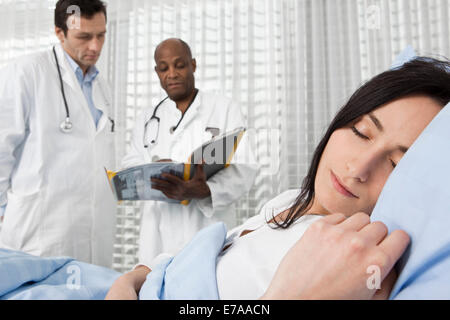 Ein Patient schläft in einem Krankenhausbett während zwei Ärzte im Hintergrund über ihr Horoskop konsultieren Stockfoto